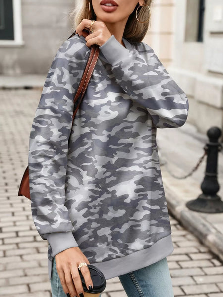 A woman wearing a gray and white camouflage sweatshirt, paired with blue jeans and holding a brown leather shoulder bag, standing on a cobblestone street.