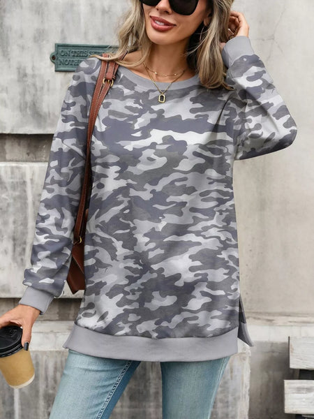 A woman wearing a gray and white camouflage long-sleeve sweatshirt, styled with light blue jeans, a brown shoulder bag, and sunglasses, standing against a textured gray wall.