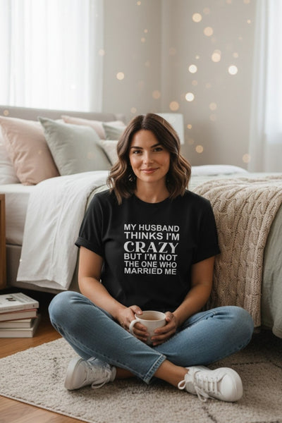 A woman sits cross-legged on a rug in a bedroom setting, wearing a black graphic t-shirt with white text that reads 'MY HUSBAND THINKS I'M CRAZY BUT I'M NOT THE ONE WHO MARRIED ME'.