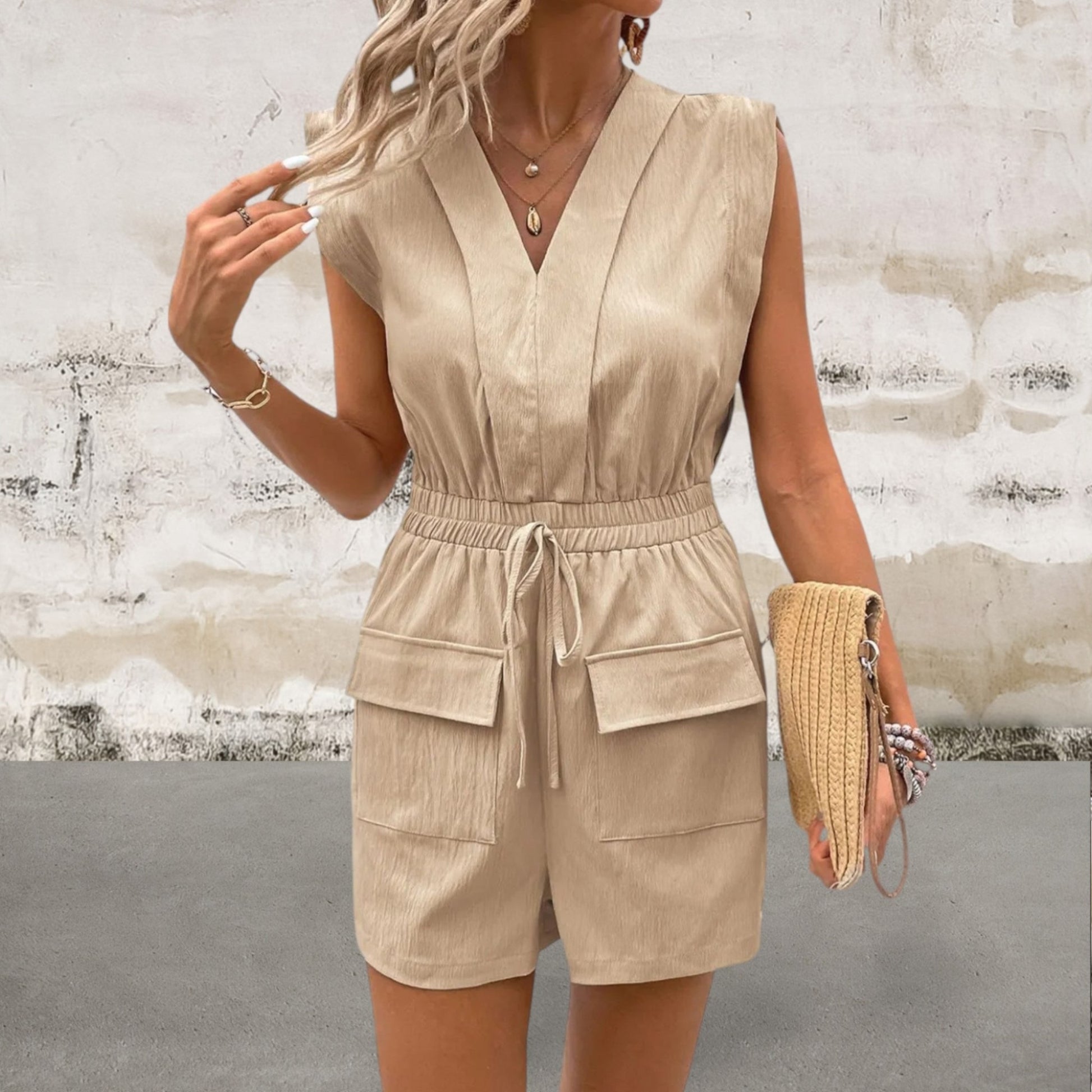 Woman wearing a beige sleeveless jumpsuit against a textured wall.